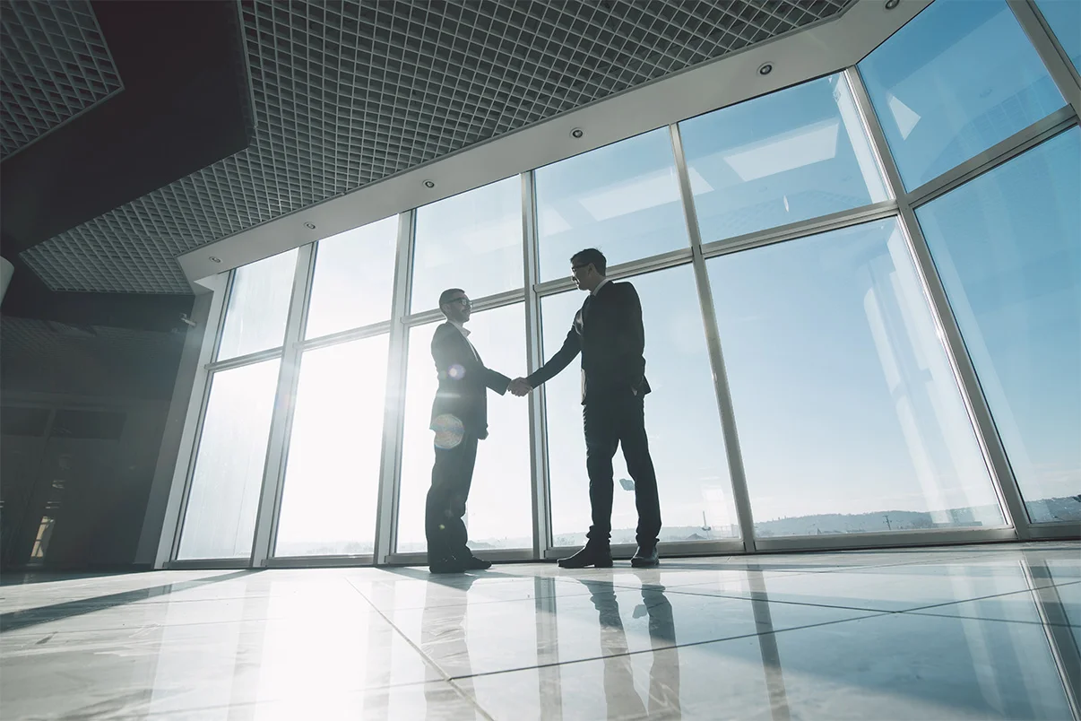 Two young businessmen are shaking hands with each other standing against panoramic windows